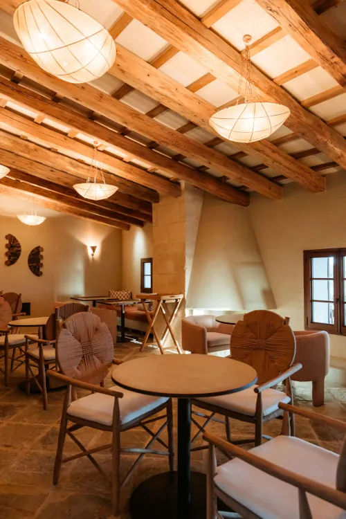 Warm, inviting café with rustic decor; features wooden beams, soft lighting from pendant lamps, round tables, and cushioned chairs against a stone wall backdrop.