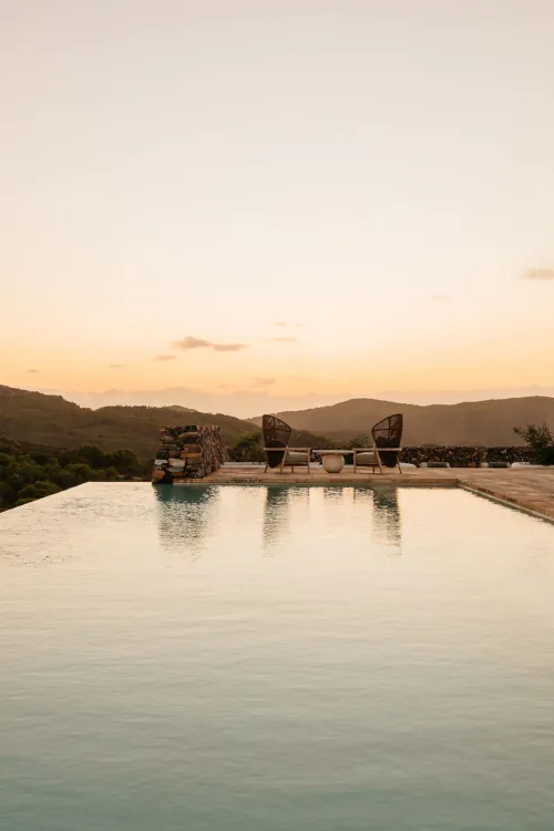 Infinity pool at sunset overlooking distant mountains. Two wicker chairs on the deck create a serene, tranquil atmosphere under the soft, warm sky.
