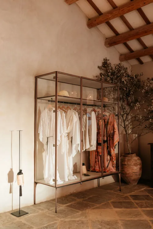 Clothing display in a glass cabinet with white and patterned garments. Rustic room with wooden beams, stone floor, and a large potted plant in the corner.