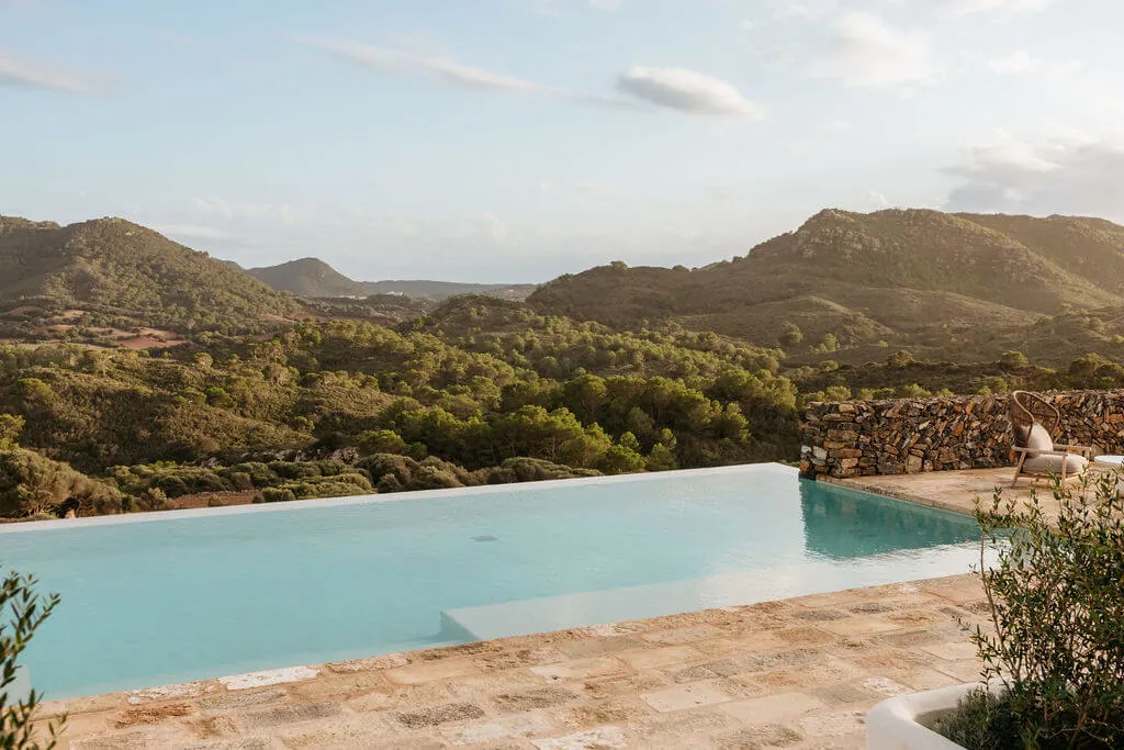 Infinity pool overlooking lush green hills under a partly cloudy sky. A stone wall and rustic chair add a serene, tranquil atmosphere.