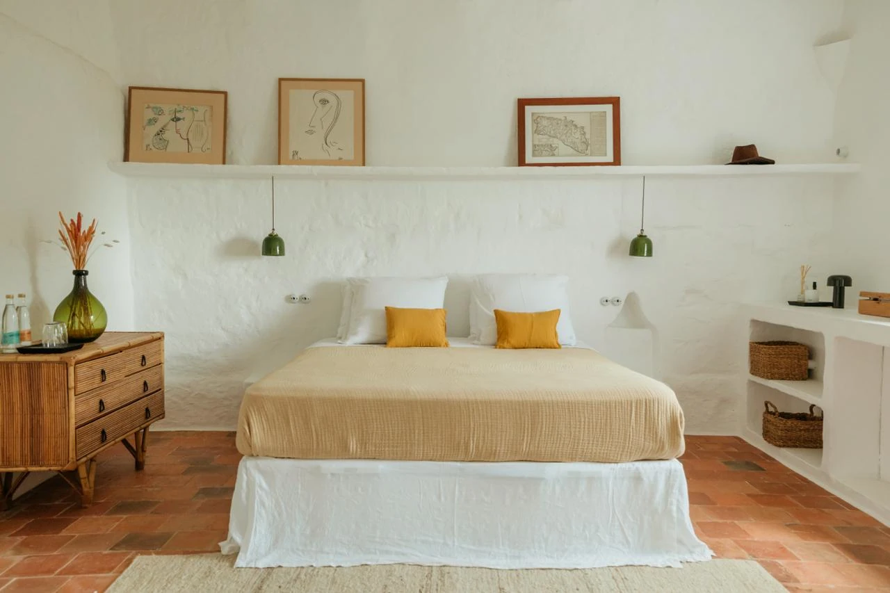 Cozy bedroom with a warm beige bed, white pillows, and yellow cushions. Green pendant lights hang above. Art on a shelf, rustic decor, earthy tones.