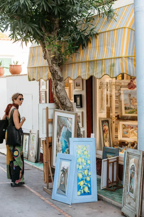 A woman in vibrant pants stands outside an art gallery, examining framed paintings displayed under a striped awning. The scene is serene and artistic.