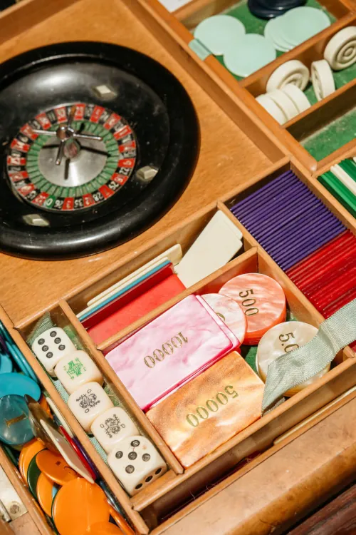 A wooden box contains a roulette wheel, colorful poker chips, dominoes, and dice, evoking a vintage casino vibe focused on gaming and chance.