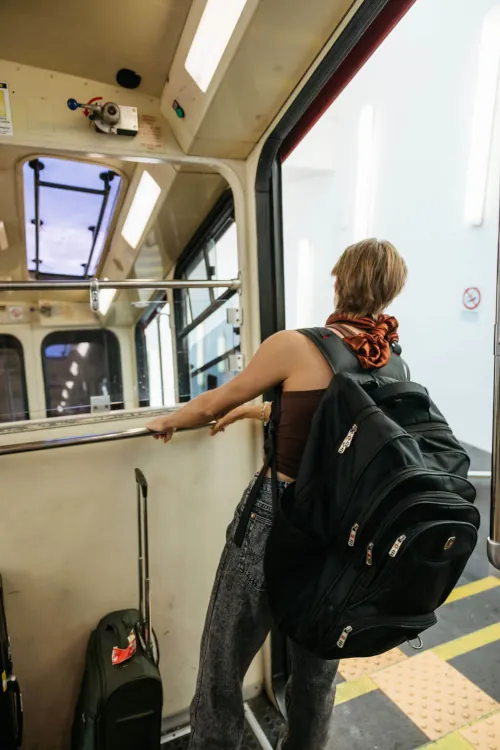 A person with short hair and large backpack stands in a tram, gazing out the open door. They hold onto a rail, alongside their suitcase. The mood is adventurous.
