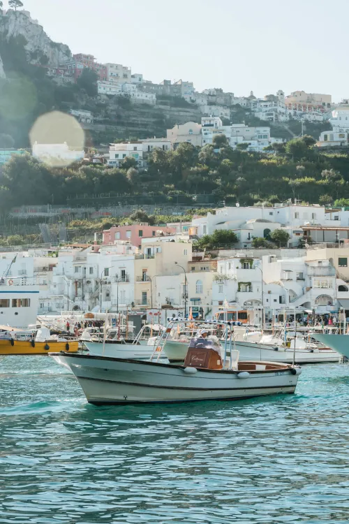 A small boat sails on turquoise water in a picturesque harbor. White and pastel houses rise up a lush hillside under a clear sky, conveying a serene, coastal vibe.