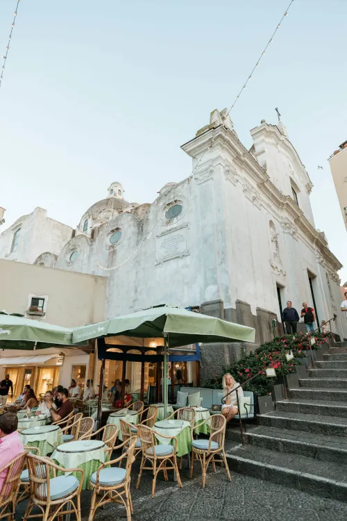 Outdoor café with green umbrellas and people dining beside an old white church. Stone steps lead upward; the mood is relaxed and inviting.