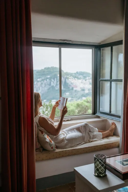 A woman in white lounges on a window seat, reading a book. The window reveals a scenic hilly view. The scene feels peaceful and cozy.