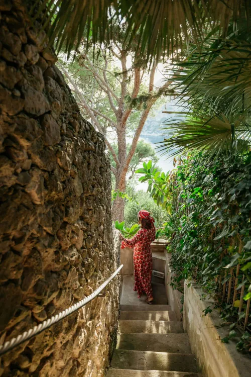 A person in a red patterned dress and hat descends a stone staircase surrounded by lush greenery, leading to a scenic view of a distant tree-filled landscape.