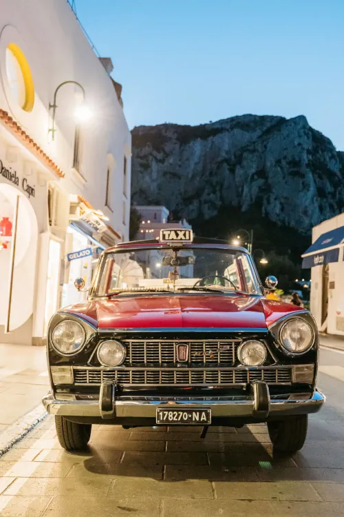 Vintage red Fiat taxi on a narrow street, flanked by quaint storefronts under a twilight sky, with a rugged mountain in the background.