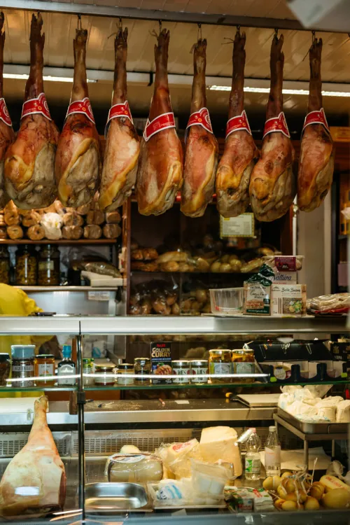 Hanging cured hams with red labels in a deli; shelves display bread, jars, cheeses, and sandwich fixings, conveying a bustling, savory atmosphere.