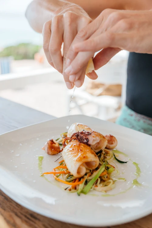 A hand squeezes lemon over grilled calamari atop a bed of colorful vegetables on a white plate, conveying freshness and Mediterranean cuisine.