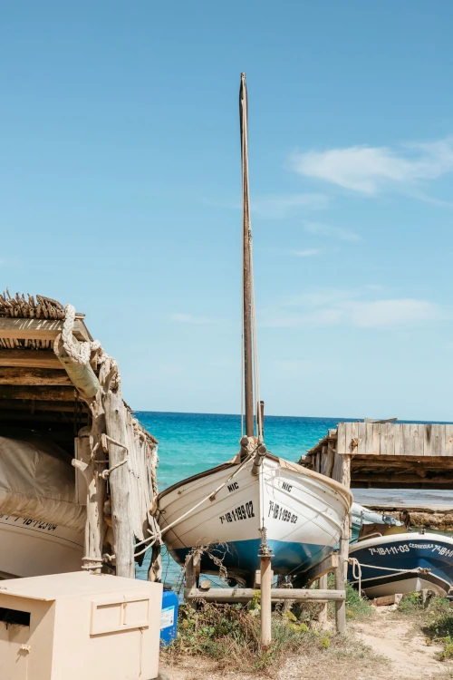 A white wooden boat sits between rustic shelters on a sandy beach. The clear blue sea and sky create a serene, coastal atmosphere.