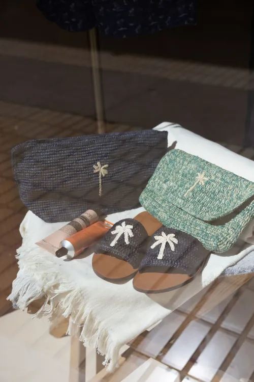 Display of woven accessories on a fringe blanket: a navy waist bag, a green purse, and sandals, each adorned with a palm tree motif, plus a tube of cosmetic cream.
