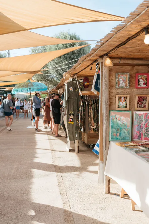 Outdoor market scene with wooden stalls displaying colorful artwork and vintage clothing, under tan sunshades. Shoppers stroll along sunlit path. Airy, vibrant feel.