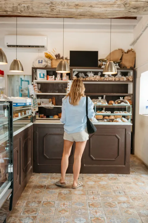 Woman in a light blouse and shorts stands at the counter of a cozy bakery, surrounded by shelves of fresh breads and pastries under warm pendant lights.