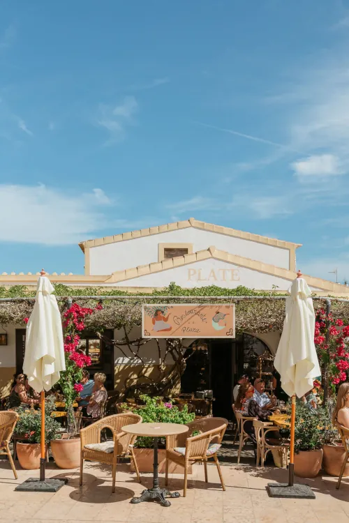 Outdoor café with wicker chairs and tables under a pergola adorned with vibrant pink flowers, set against a clear blue sky. Relaxed and inviting atmosphere.