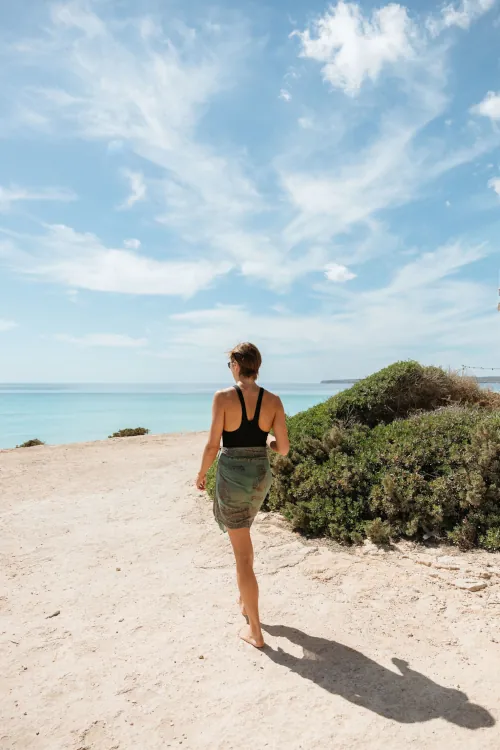 A woman in a black tank top and green skirt walks barefoot on a sandy beach, with lush bushes nearby and a calm blue sea under a partly cloudy sky.