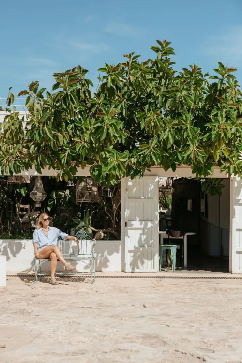 A woman sits on a white bench outside a rustic building with open doors, surrounded by lush green foliage under a clear blue sky, conveying tranquility.