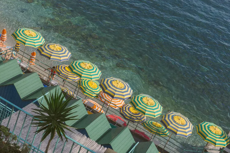 Aerial view of a vibrant beach scene with yellow and green striped umbrellas lining the shore, clear turquoise water, and green-roofed cabins. Relaxing vibe.