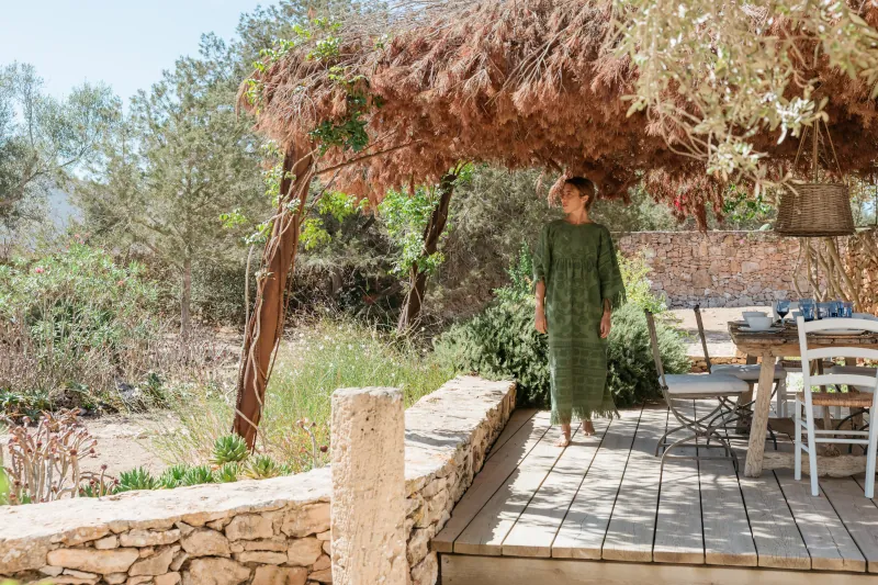 A woman in a green dress stands on a rustic wooden deck shaded by a thatched roof, surrounded by lush greenery and rustic stone walls, evoking tranquility.