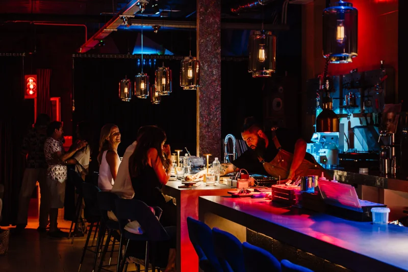 Dimly lit bar with blue and red lighting, patrons seated at the counter. A bartender prepares drinks, creating an intimate and lively atmosphere.