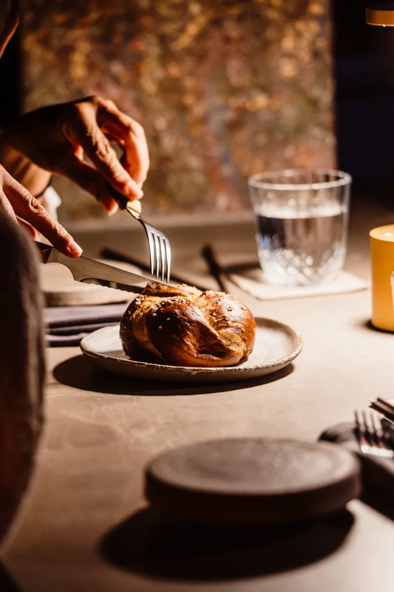 A hand slices a rustic loaf on a ceramic plate under warm light. Beside it, a glass of water and a yellow cup sit on a table, evoking a cozy ambiance.