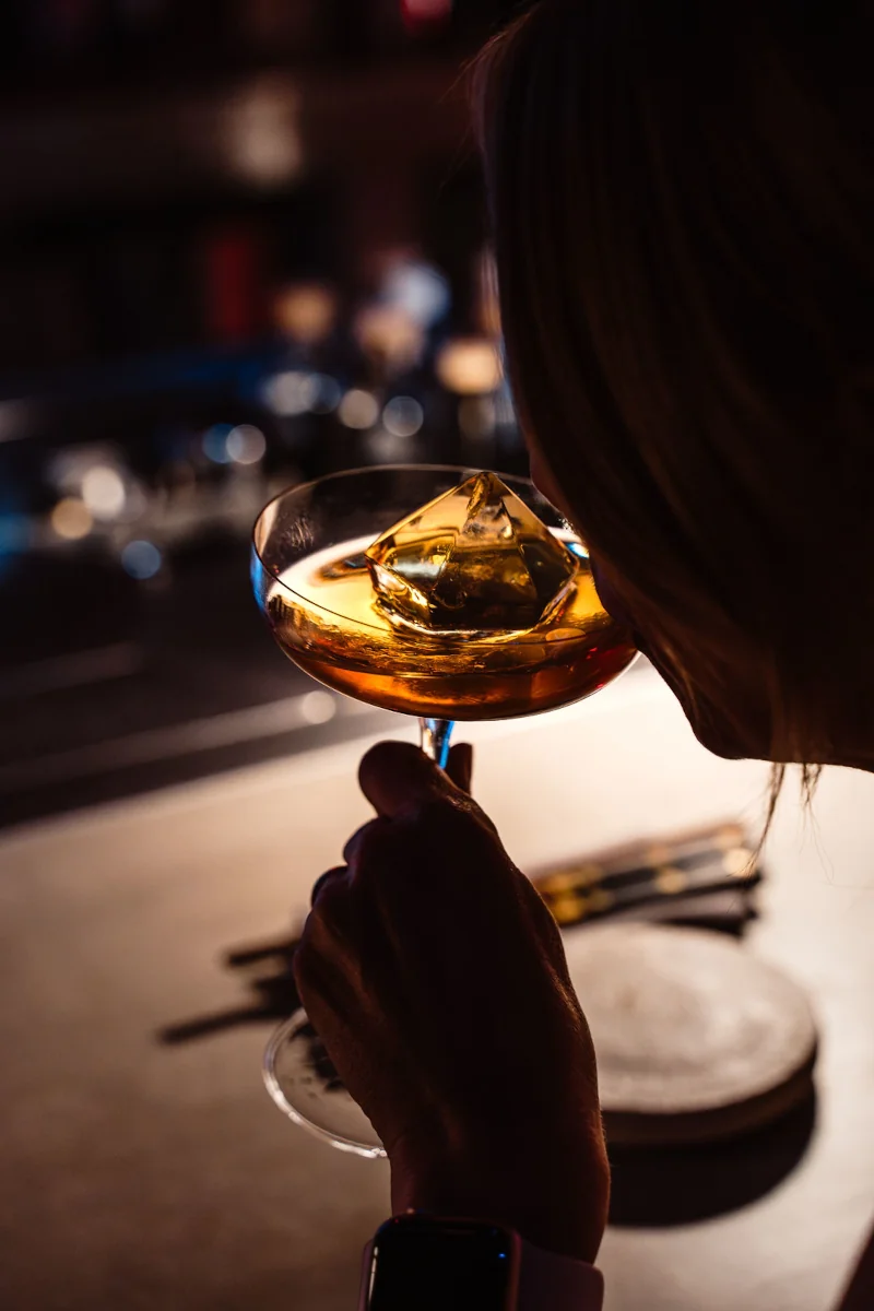 A person sipping an amber cocktail with a large ice cube in a dimly lit bar. The atmosphere is warm and intimate, with soft focus in the background.