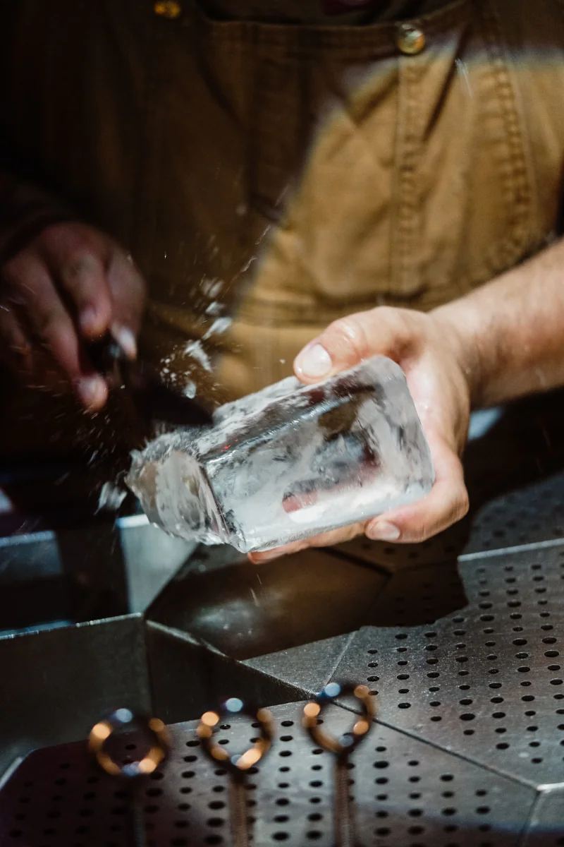 A person in an apron carves a clear ice block. Water splashes around, capturing movement and focus. The scene conveys craftsmanship and precision.