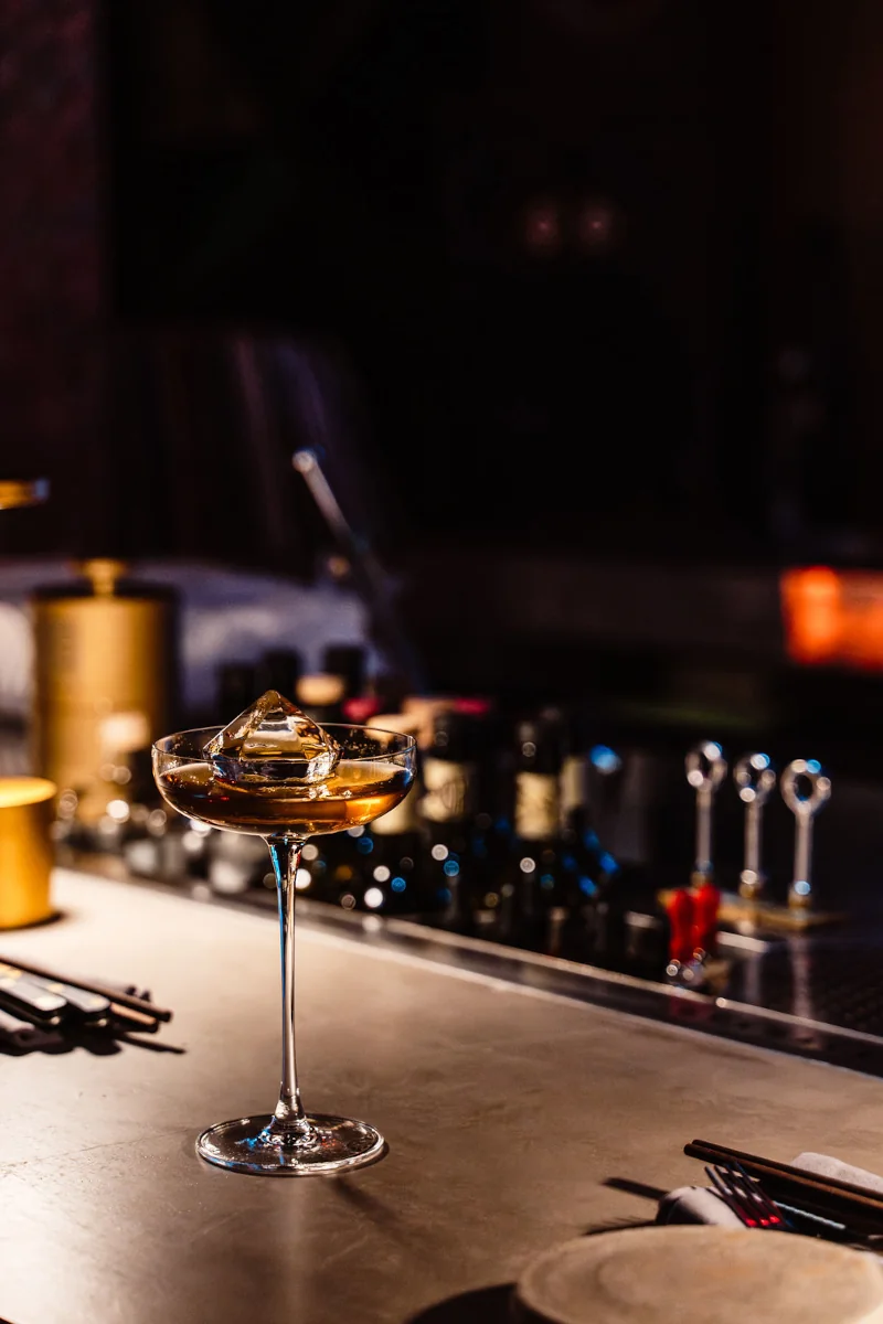 A cocktail glass with amber liquid and a large ice cube sits on a dimly lit bar counter. In the background, bottles and bar tools create an elegant ambiance.