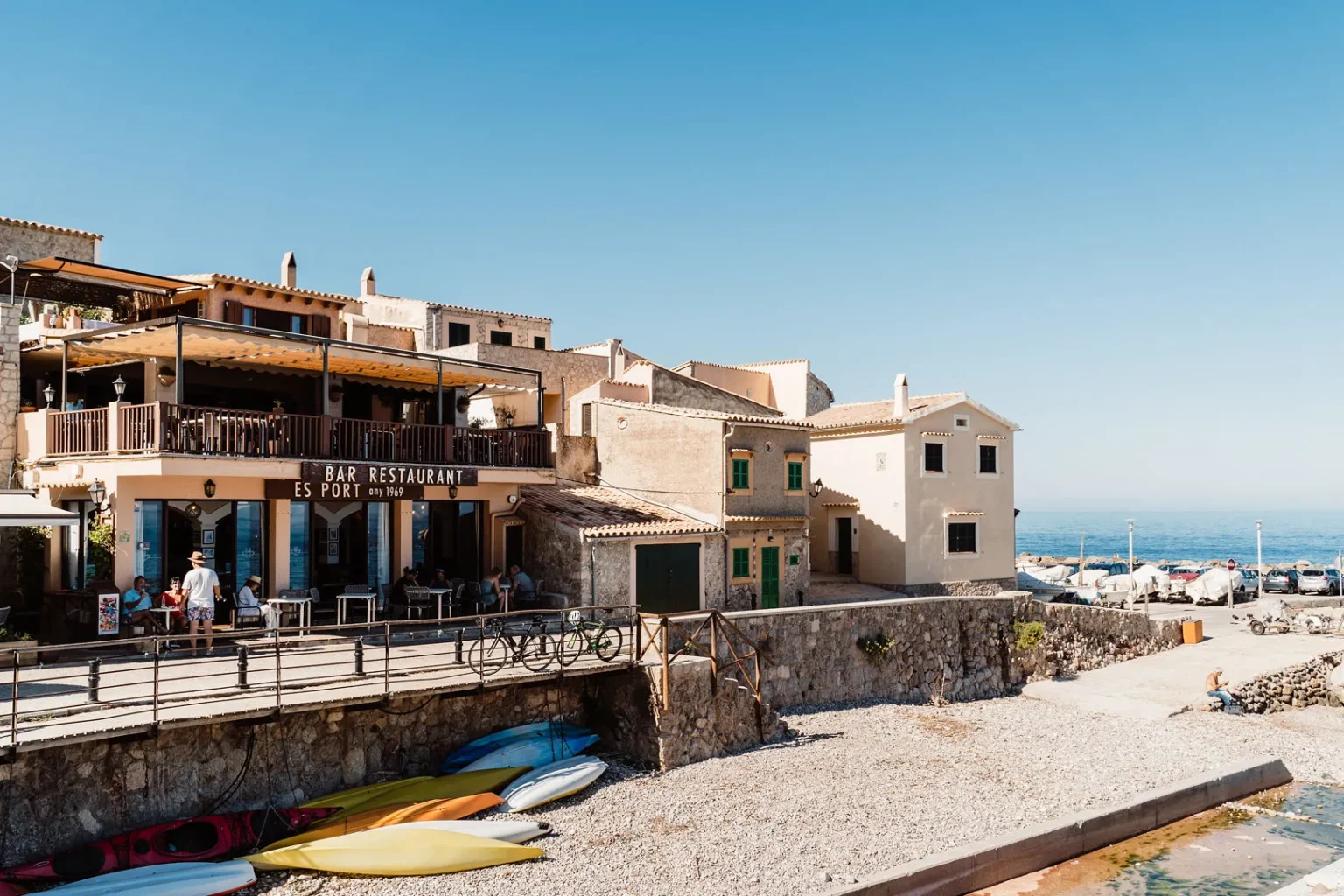 Coastal scene featuring a rustic bar and restaurant with people dining on a terrace. Colorful kayaks lie on a stone beach below, with the ocean beyond.