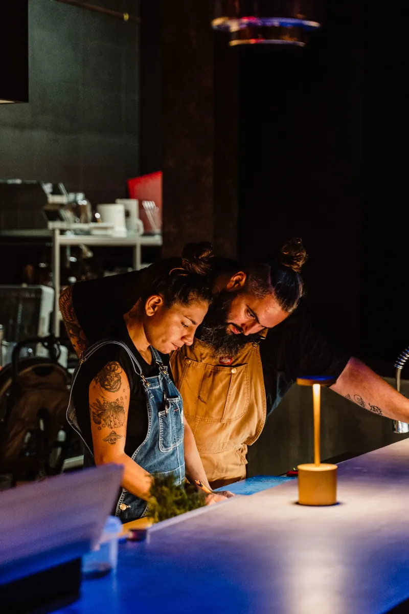 A man and woman, both in aprons, closely examine something on a table in a dimly lit kitchen. They appear focused and engaged in their task.
