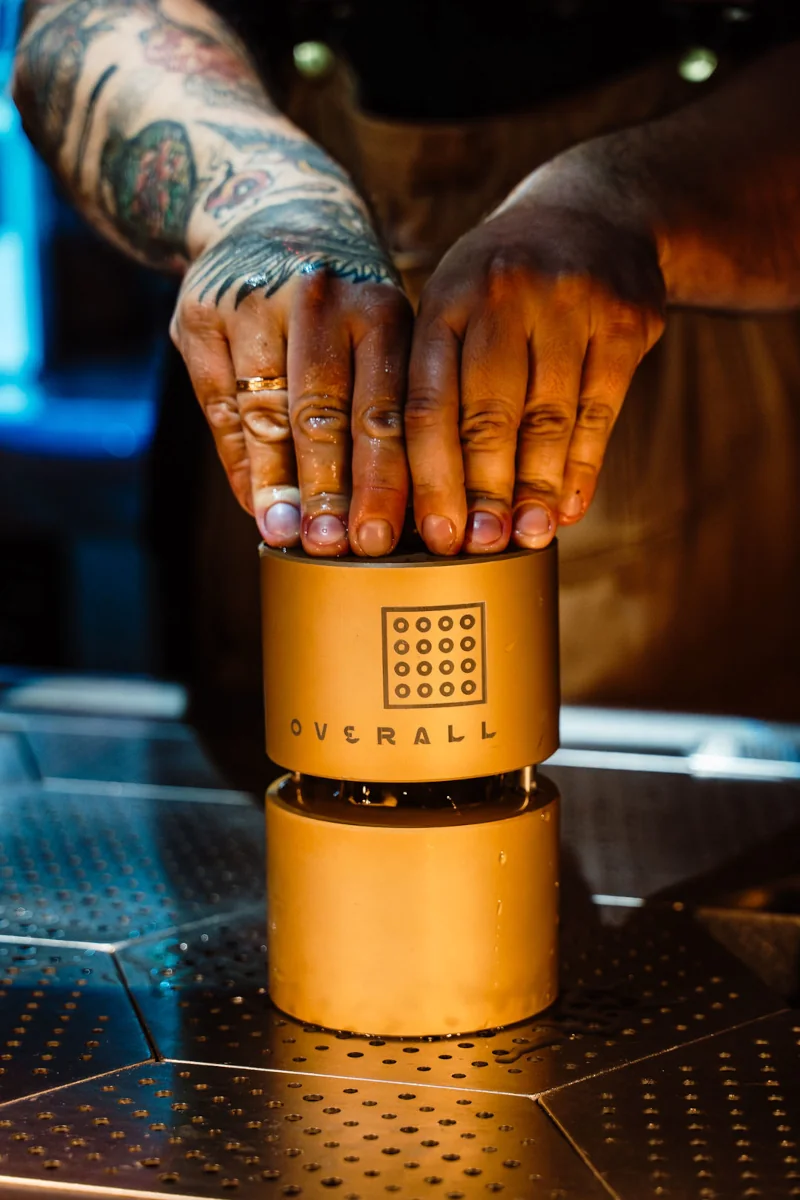 Hands with tattoos pressing down on a bronze coffee tamper labeled 
