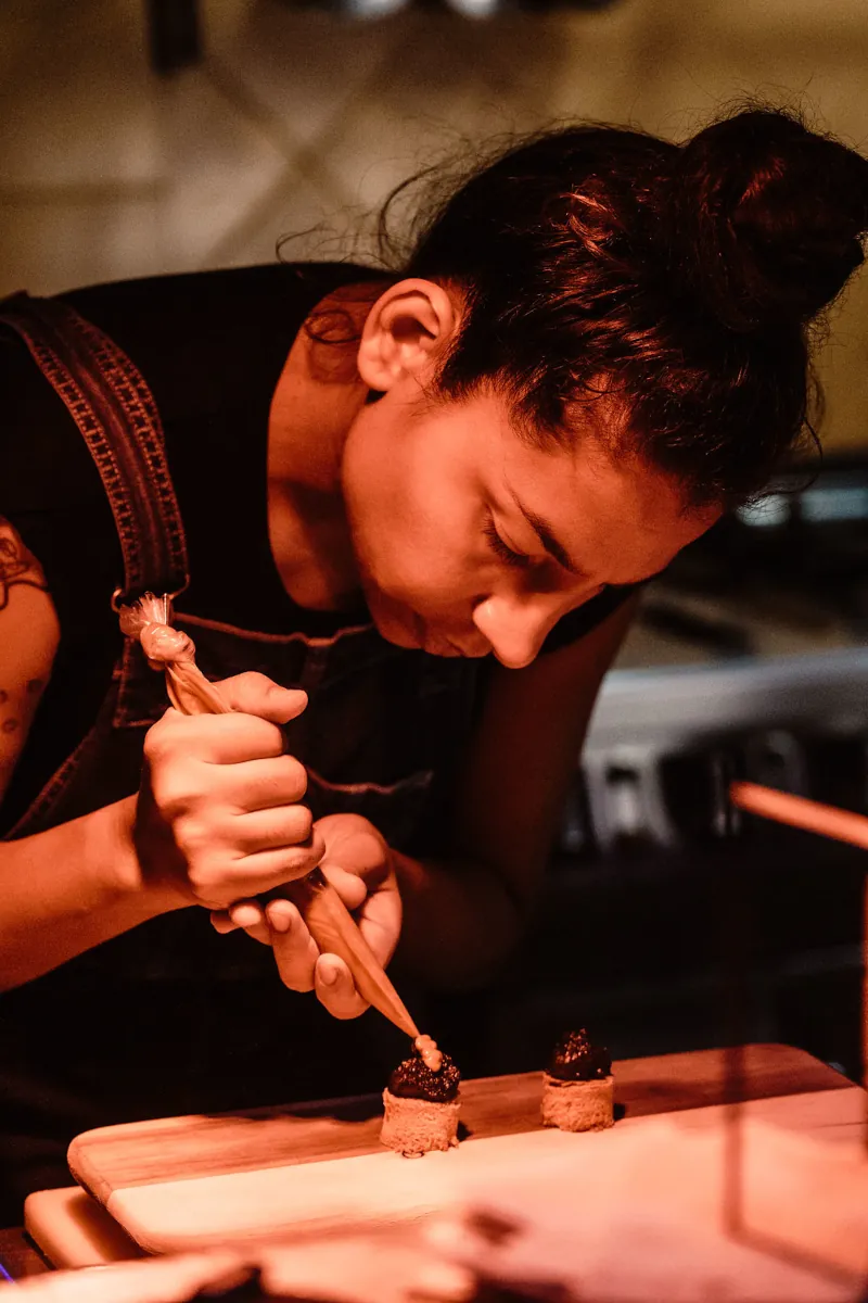 A focused chef in a dimly lit kitchen decorates small, round desserts on a wooden board with a pastry bag. The atmosphere is warm and detailed-oriented.