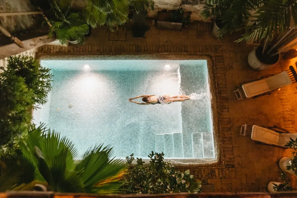 A person swims leisurely in a small, illuminated outdoor pool, surrounded by lush green plants and cozy deck chairs, creating a relaxing atmosphere.