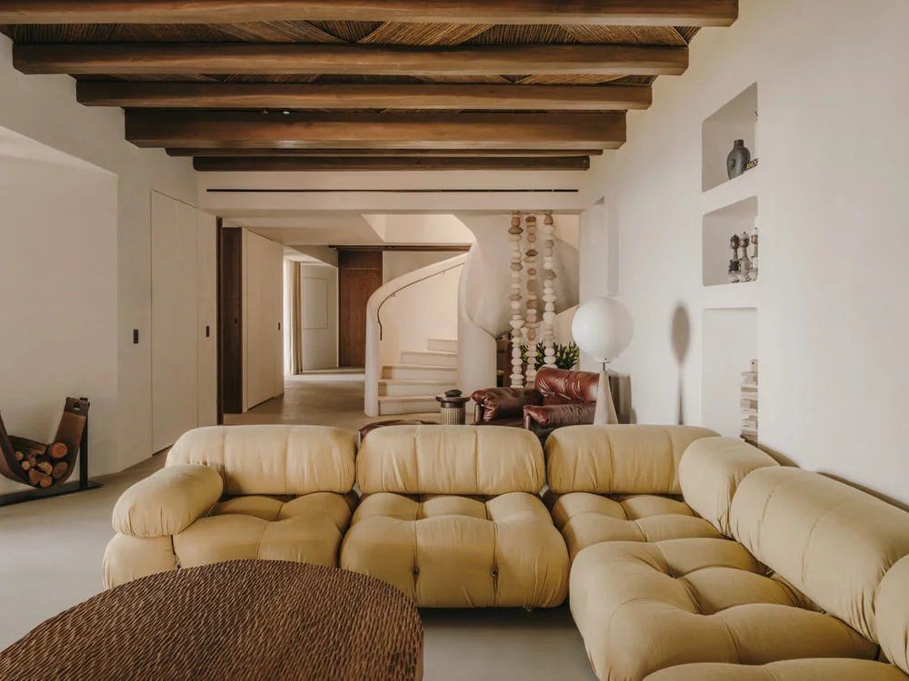 Spacious living room with beige tufted sofa, rustic wooden beams, woven coffee table, and minimalist decor. A spiral staircase adds elegance. Cozy ambiance.