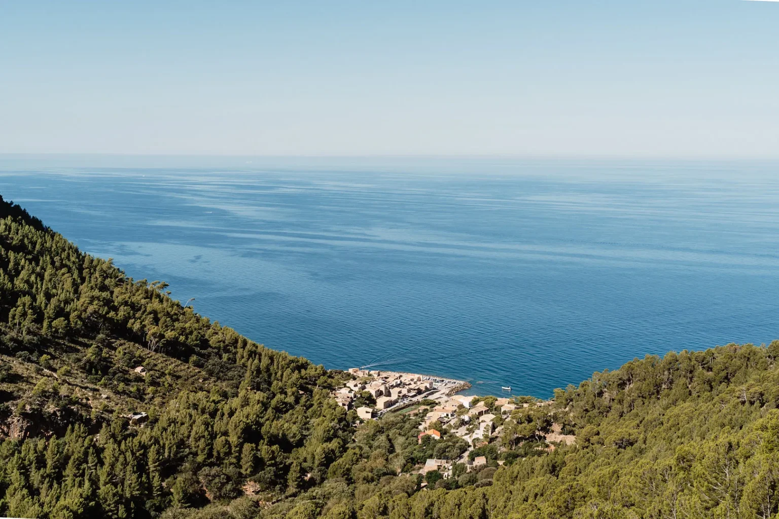 Expansive view of a calm, blue sea under a clear sky. Green, forested hills frame a coastal village with scattered houses, evoking tranquility and peace.