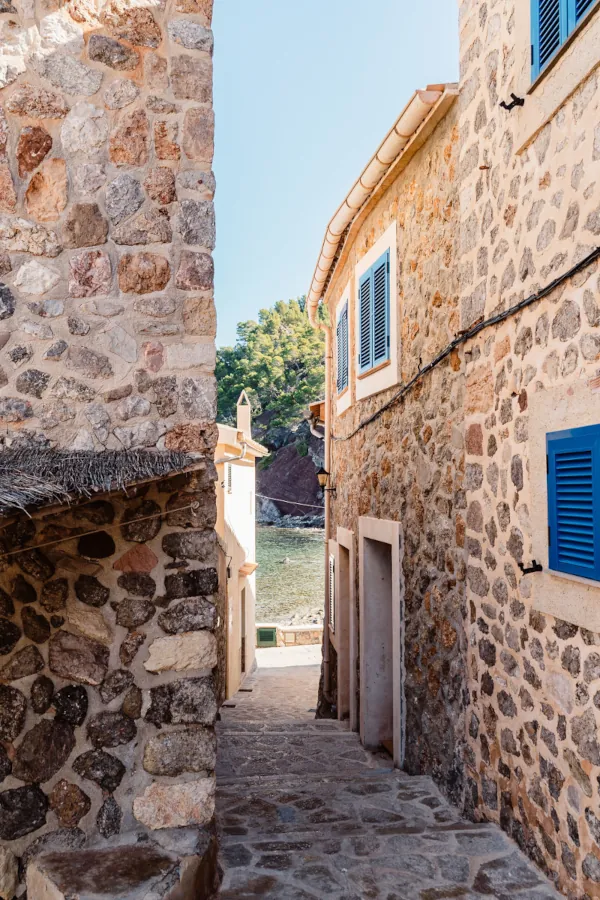 Narrow stone alley between rustic buildings with blue shutters, leading to a view of the sea and distant green hills under a clear sky. Tranquil mood.