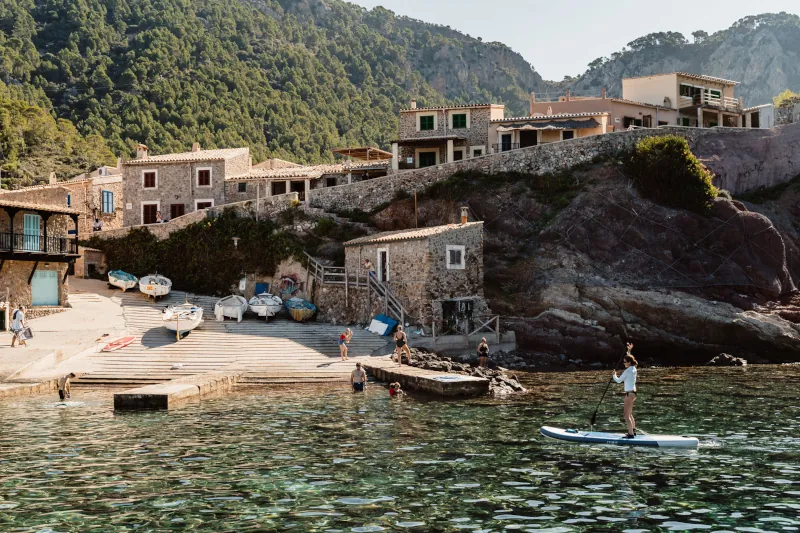 A serene coastal village nestled against lush hills, with rustic stone buildings. People swim and paddleboard in clear blue water, creating a peaceful scene.