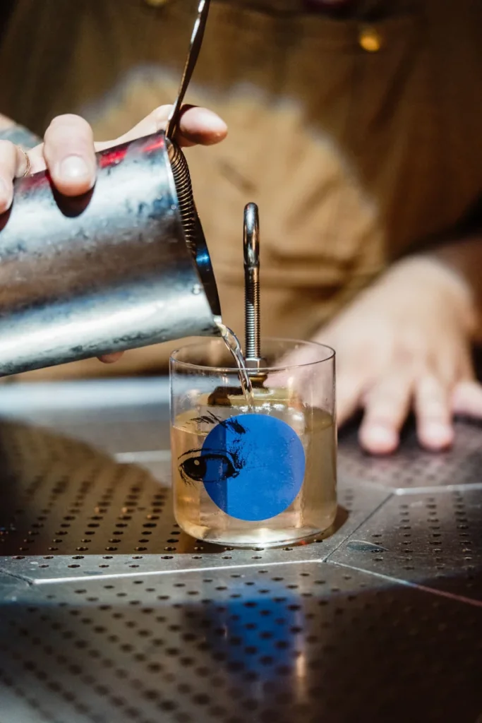 A bartender pours a drink through a strainer into a glass with a blue circle and eye design. The scene is lit warmly, suggesting a cozy bar atmosphere.