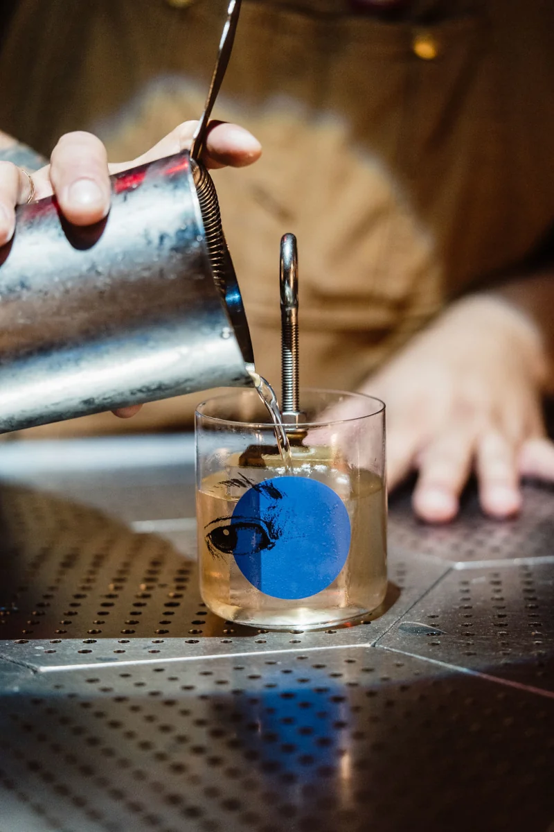 A bartender pours a drink through a strainer into a glass with a blue circle and eye design. The scene is lit warmly, suggesting a cozy bar atmosphere.
