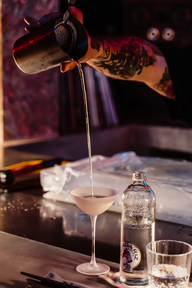 A tattooed arm gracefully pours a clear cocktail into a glass from a shaker. The setting is a dimly lit bar with a bottle and a glass in the foreground.