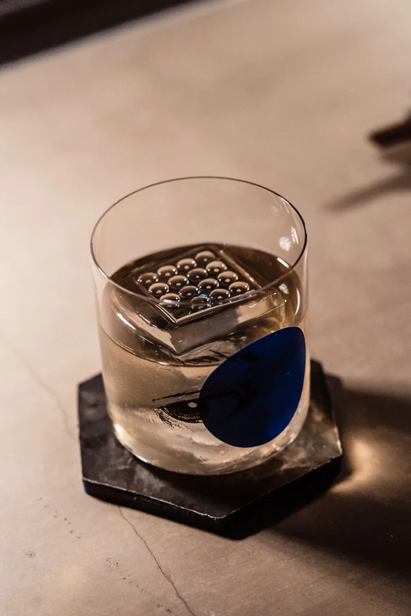 A clear glass filled with light-colored liquid and a decorative square ice cube with bubbles on a black hexagonal coaster on a smooth table.