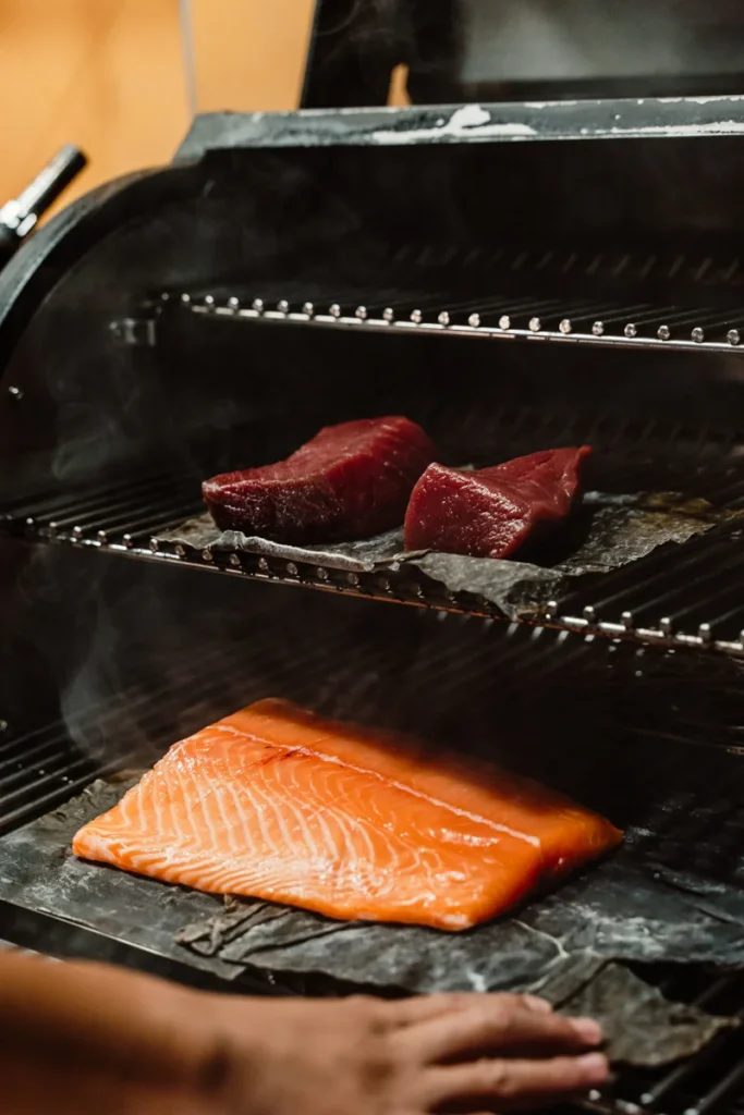 Two slabs of red meat and a salmon filet are being smoked on black slate inside a grill. An open hand is adjusting the slate, with steam visible.