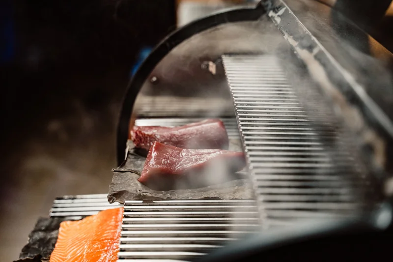 Close-up of a grill with two red tuna steaks and an orange salmon fillet cooking, surrounded by rising steam, creating a smoky, appetizing scene.