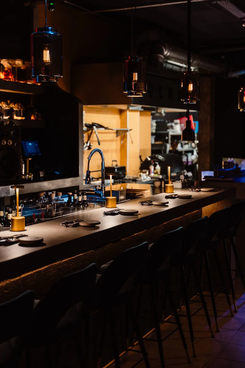 Dimly lit modern bar interior with a sleek countertop, tall chairs, hanging lights, and a visible open kitchen, creating a cozy, inviting ambiance.