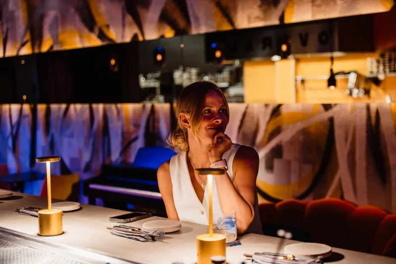 A woman sits at a bar with a thoughtful expression, surrounded by soft, ambient lighting. The setting is modern and artistic, with abstract wall designs.