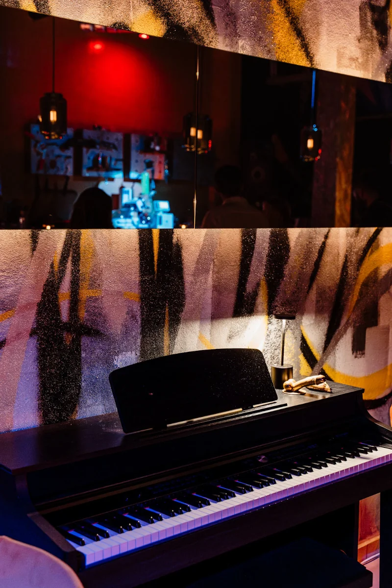 Electric piano in dimly lit room with graffiti walls. A mirror reflects a vibrant bar with red lights and bottles, creating a moody, artistic ambiance.