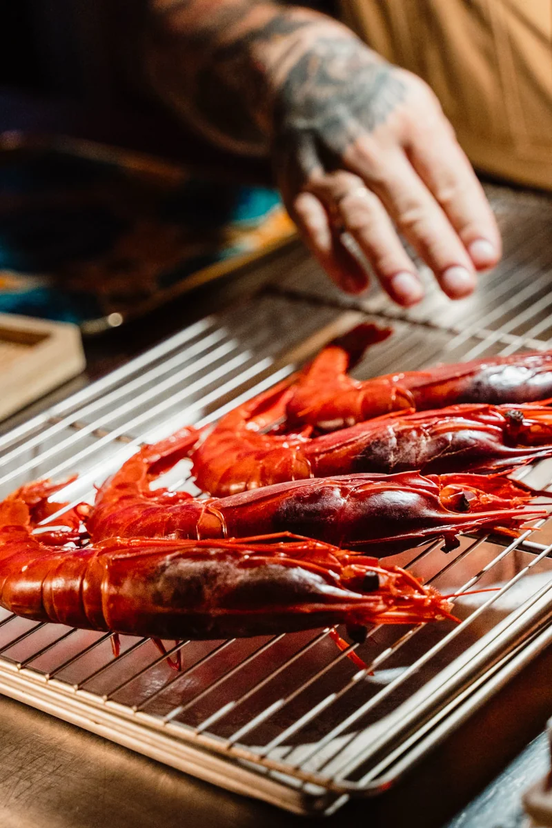 A tattooed hand hovers over vibrant red lobster tails resting on a metal rack. The scene suggests cooking preparation, evoking a sense of culinary anticipation.