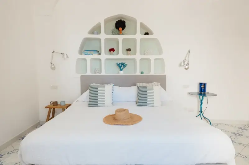 Spacious bedroom with a white bed, two striped pillows, and a straw hat. Modern wall shelving with decor accents; minimalistic and serene vibe.