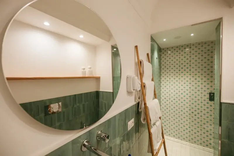 Modern bathroom with green tiles, round mirror, wooden towel ladder, and patterned shower area. Brightly lit, creating a clean, calming ambiance.